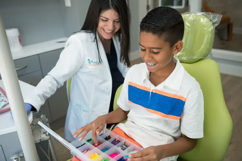 Smiling boy with braces selecting colorful orthodontic rubber bands, assisted by a board-certified orthodontist in a modern Miami orthodontics office.