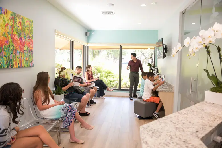 Modern orthodontic waiting area at Miami Orthodontic Specialists featuring patients relaxing with magazines, children using tablets, and a vibrant floral artwork on the wall, emphasizing a welcoming environment for braces and Invisalign treatments.