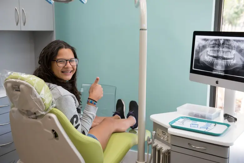 Young girl in orthodontic chair, smiling and giving a thumbs up, showcasing braces in Miami orthodontic office with dental equipment and x-ray monitor in background.